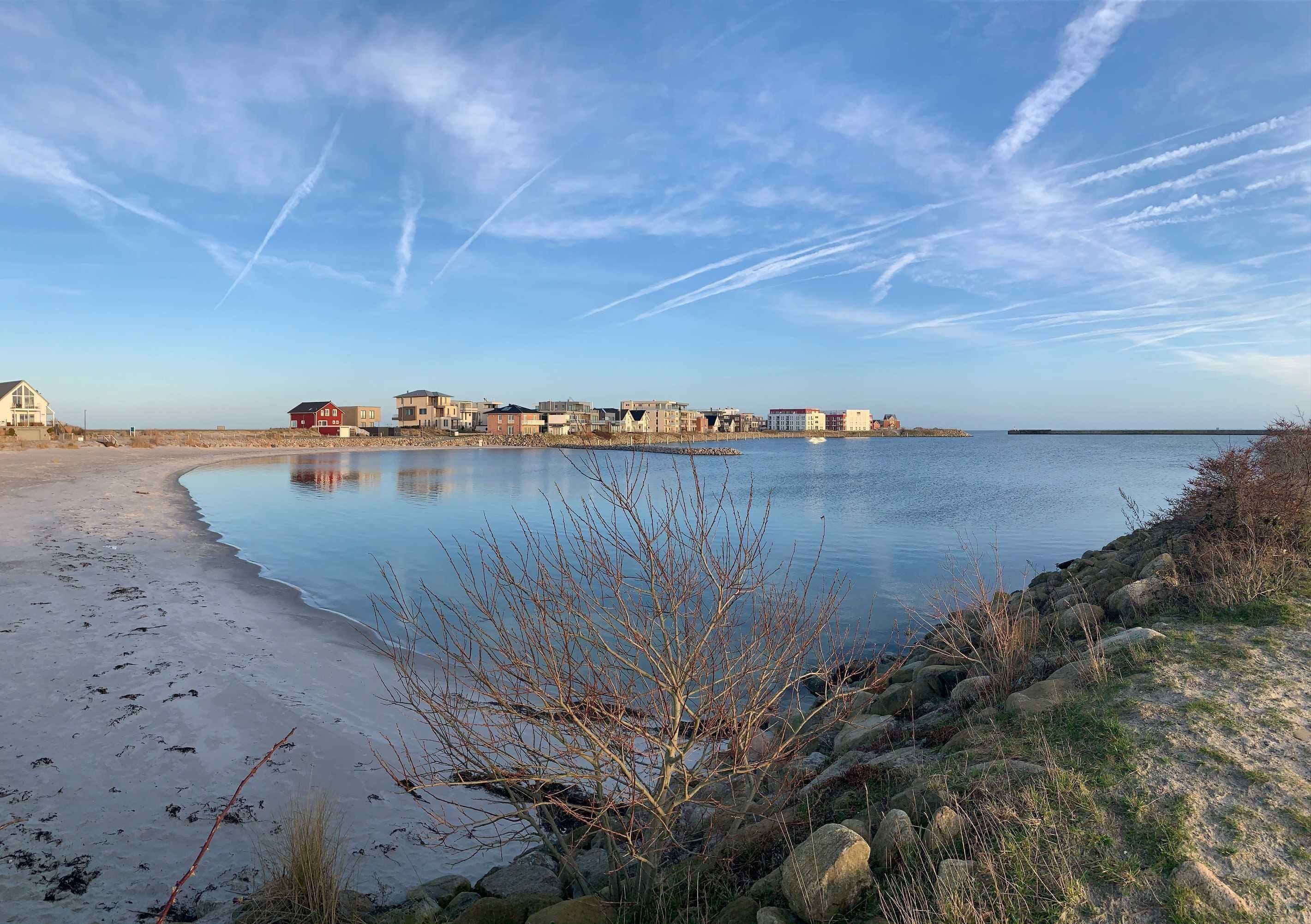 A coastal area with a sandy beach, a calm body of water, and a row of buildings in the background.