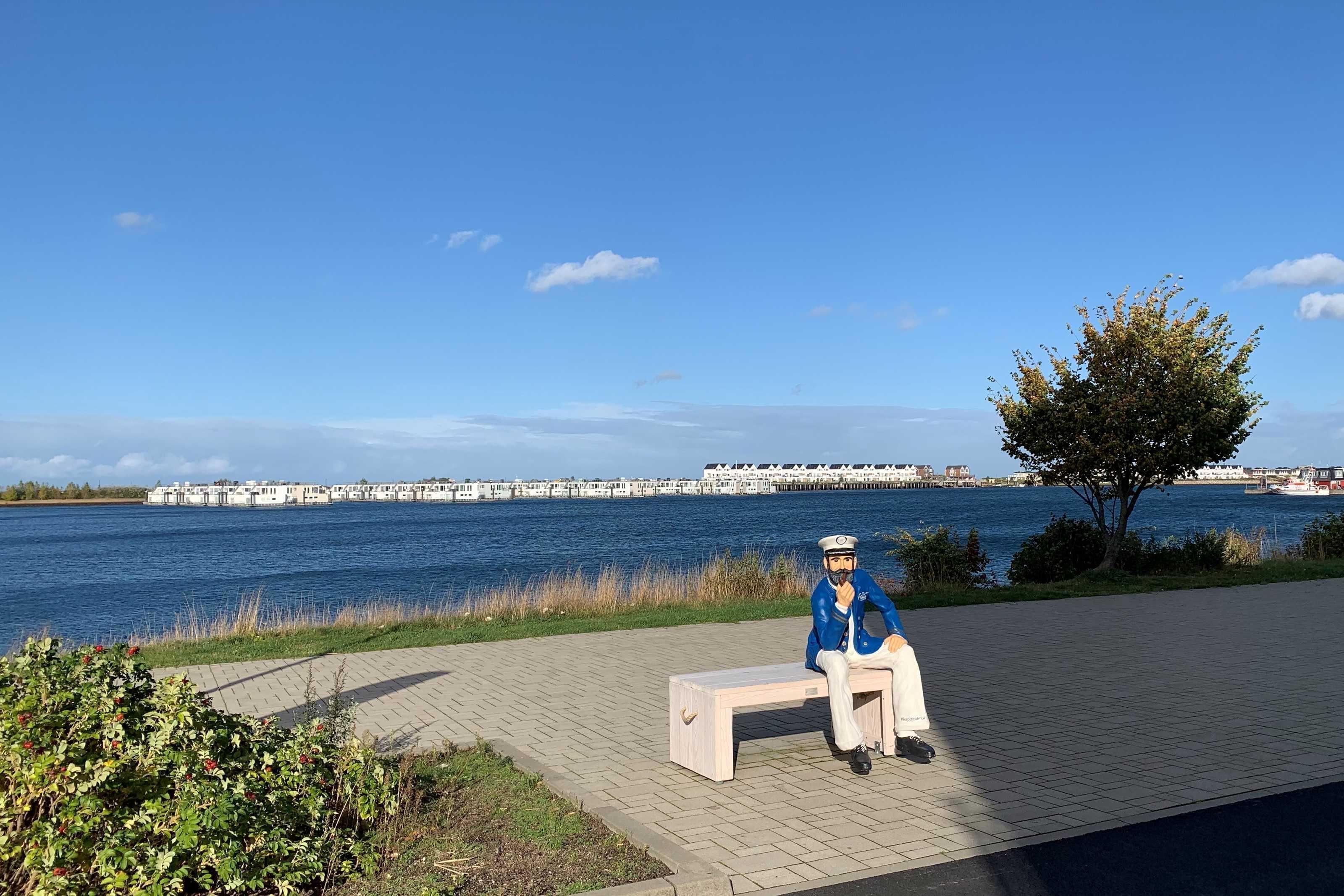 Une personne est assise sur un banc au bord de l’eau, d’où elle a une vue dégagée sur une jetée et des bateaux en arrière-plan.