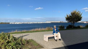 Una persona sentada en un banco frente al agua, con vista despejada a un muelle y barcos al fondo.