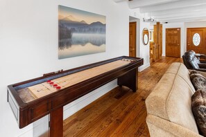A room with a shuffleboard table, a couch, and a painting of a mountain landscape. - Central A/C! HOT TUB! Play on an Acre! Near Bear Mountain/Zoo/Golf/Hiking! EV Charger! Multi-Game Tables! (Big Bear City)