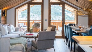 A cozy, well-lit room with a high ceiling, large windows offering a mountain view, a dining area with blue chairs and a wooden table, and a comfortable seating area with a sofa and armchairs. - 4-bedroom duplex in Méribel, near center and slopes, with concierge services (Méribel)