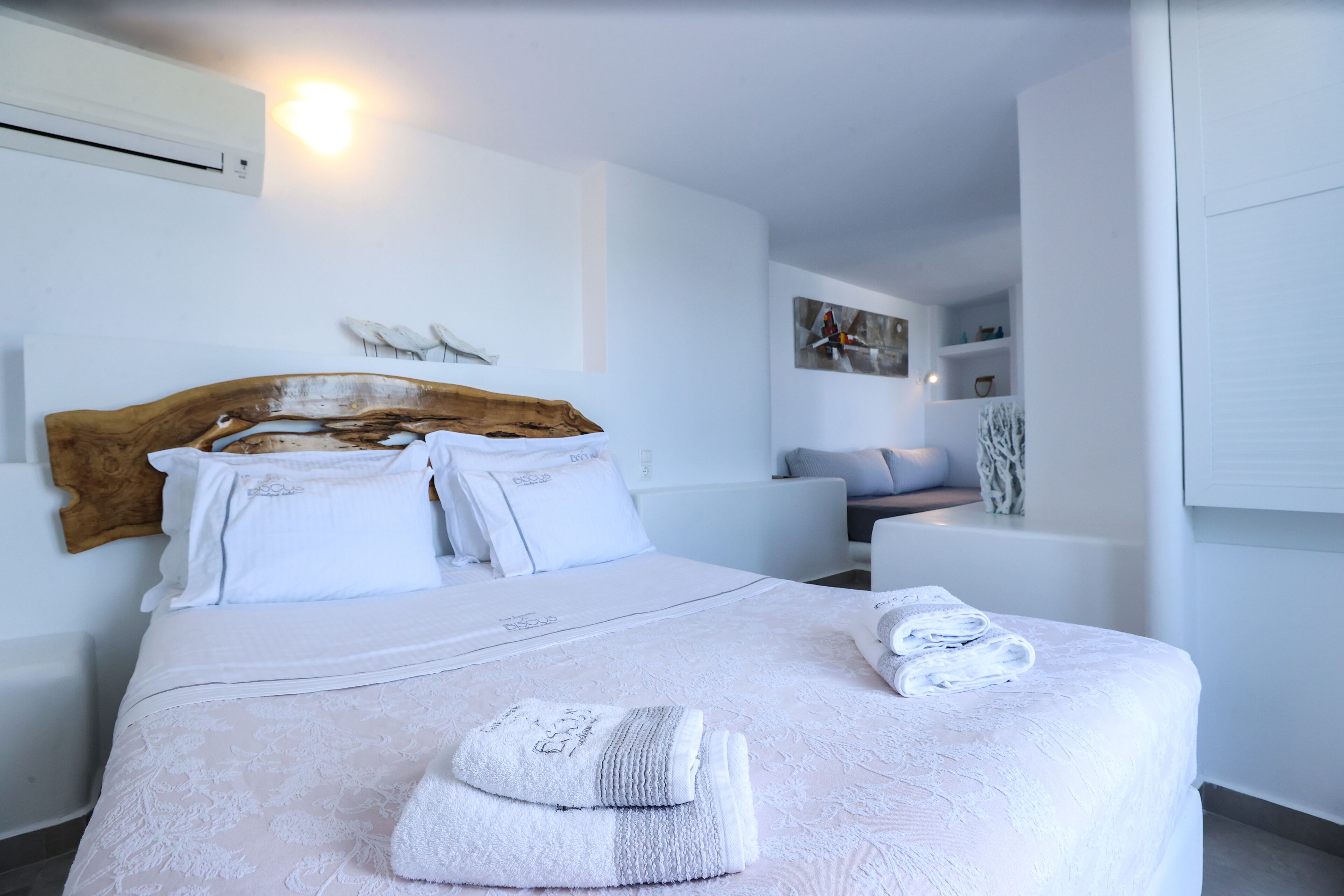 A neatly made bed with white linens and a wooden headboard, a bedside table with a vase, and a wall-mounted air conditioner.