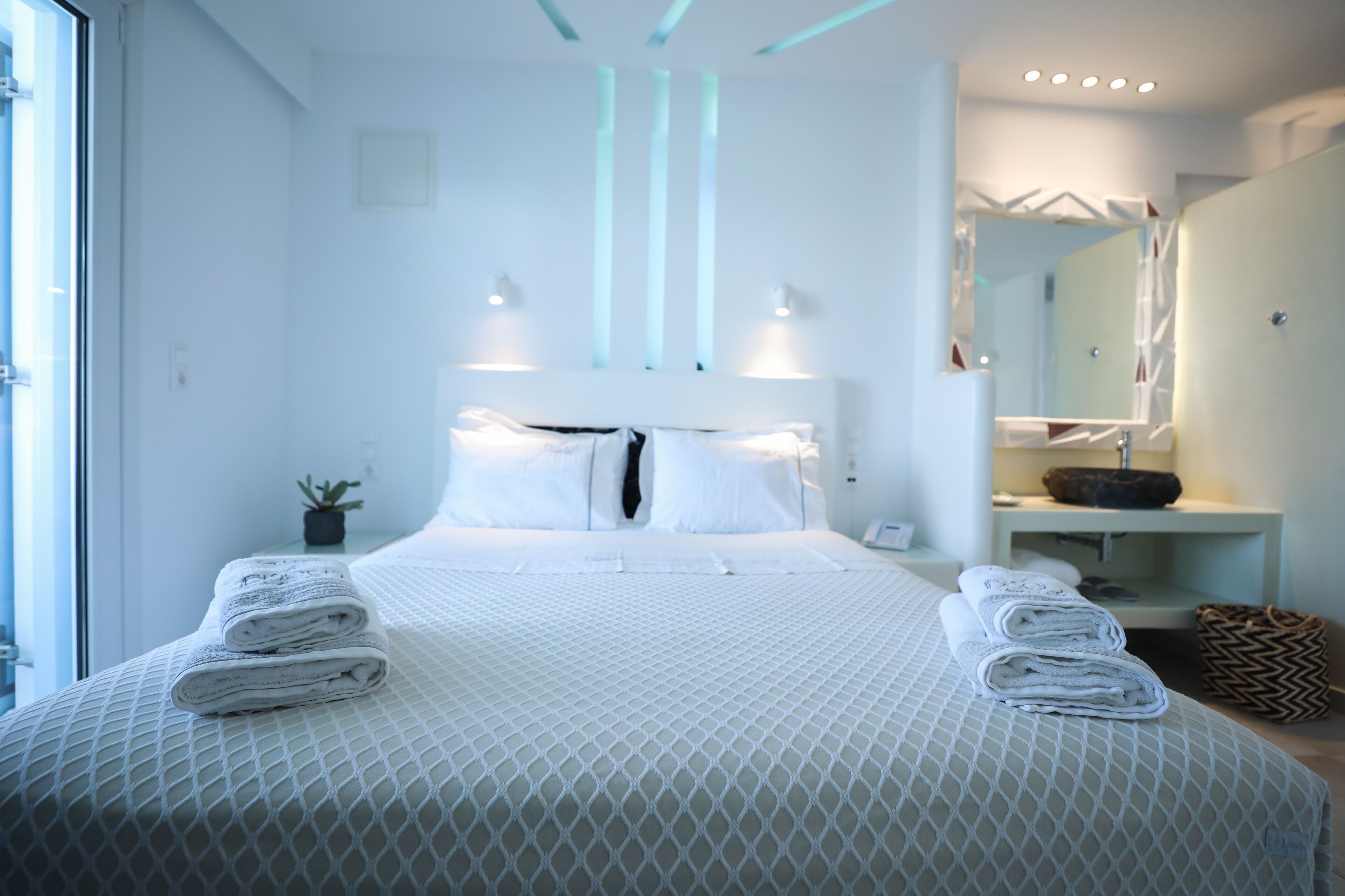 A modern hotel room with a neatly made bed, a small potted plant, and a bathroom with a mirror and sink.
