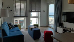 A modern living room with a blue sofa, a red ottoman, and a television. - BOW-2.15, 4 pers., 55-65sqm (HT) - Residence Bollwark 2.1 (Kappeln)