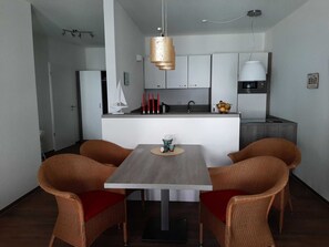 A modern kitchen with white cabinets, a central island, and a dining area with wicker chairs and a wooden table. - BOW-2.15, 4 pers., 55-65sqm (HT) - Residence Bollwark 2.1 (Kappeln)