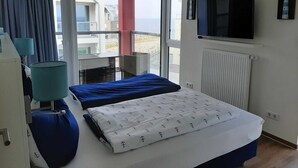 A hotel room with two beds, a TV, and a balcony with a view of buildings and the sea. - BOW-2.15, 4 pers., 55-65sqm (HT) - Residence Bollwark 2.1 (Kappeln)