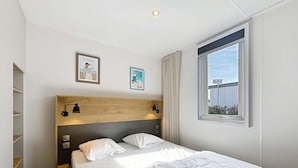 A hotel room with a bed, wooden headboard, two wall-mounted lamps, a window with curtains, and a view of a building outside.
