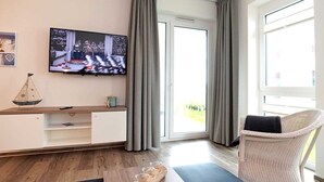 A modern living room with a flat-screen TV, a wooden console table, a wicker chair, and a large window with curtains. - BOW-2.02, 4 pers., 55-65sqm (H) - Residence Bollwark 2.0 (Kappeln)
