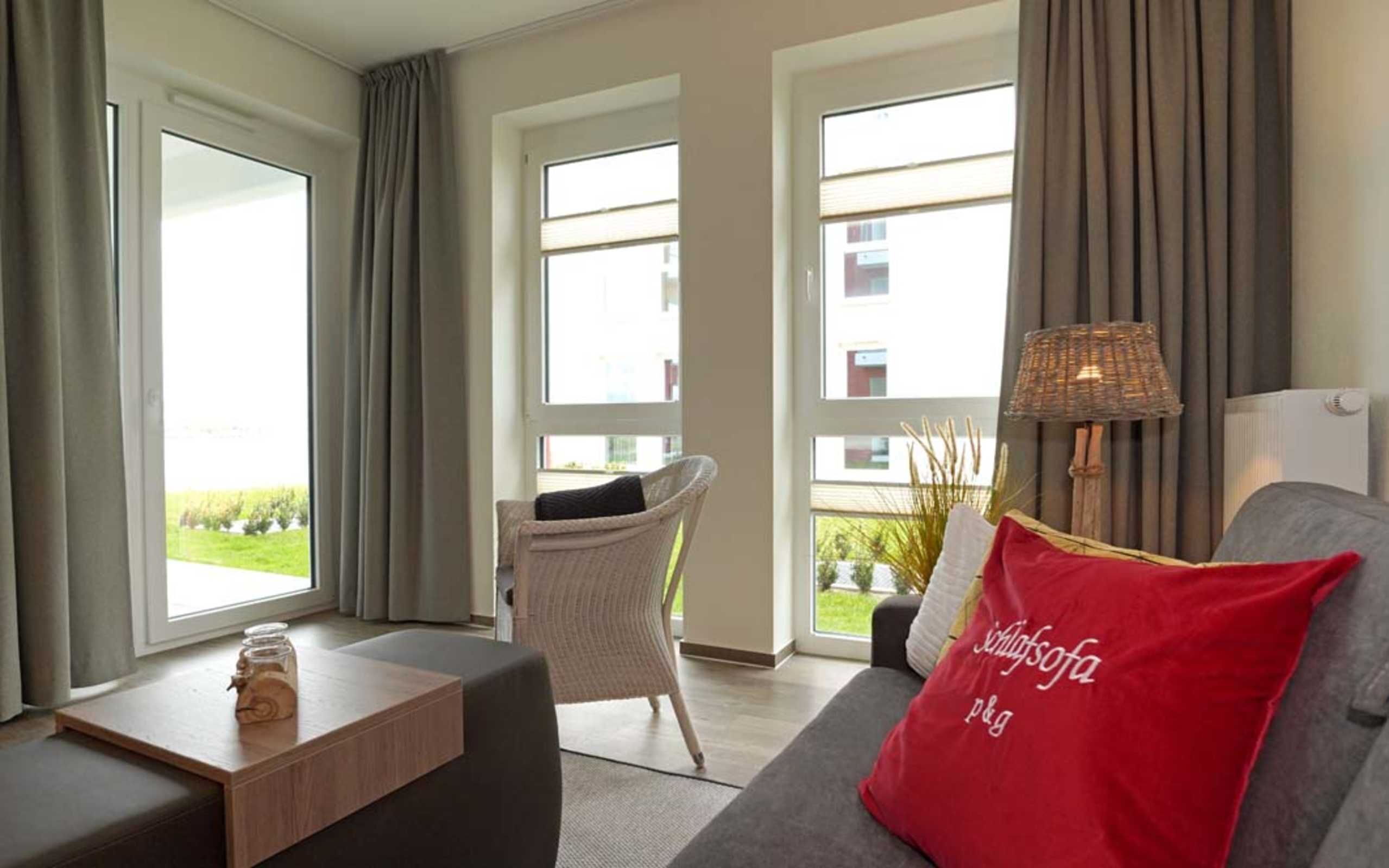A modern living room with a grey sofa, a wicker chair, and a small wooden table. There is a red pillow with text on it, and a lamp on a side table.