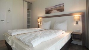A hotel room with a large bed, two bedside tables, a wall-mounted shelf, and a painting above the bed. - BOW-2.03, 4 pers., 55-65sqm (H) - Residence Bollwark 2.0 (Kappeln)