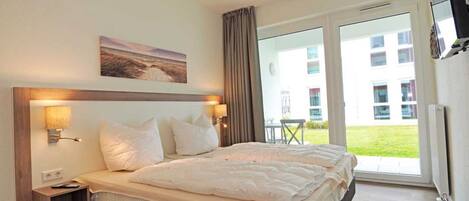 Habitación de hotel con una cama grande, una mesita de noche, un televisor montado en la pared y un ventanal corredero que da a un espacio exterior.