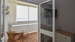En balkong med flätade stolar och ett litet bord, en skjutdörr i glas som leder in till ett rum med en TV och ett träskåp.
