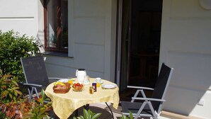 An outdoor dining area with a table set for two, surrounded by chairs, and a window with a view of greenery.