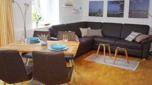 A modern living room with a dark grey sofa, a wooden dining table, and a glass coffee table. There are three framed pictures on the wall, a floor lamp, and a window with curtains.