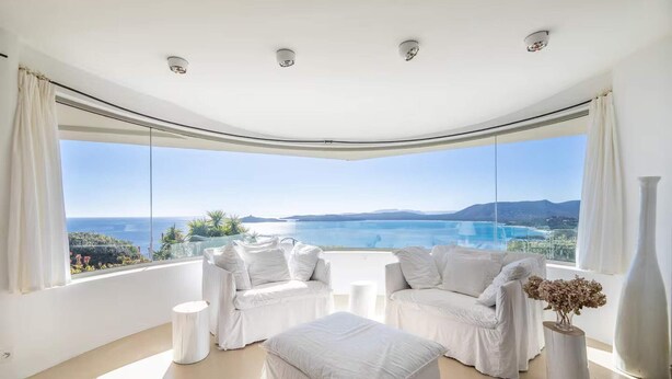 A modern room with a curved ceiling, white furniture, and a large window offering a view of the sea and mountains.
