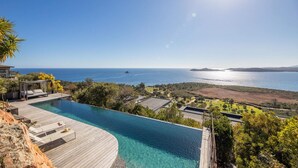 A luxurious infinity pool with a wooden deck, overlooking a coastal landscape with a clear view of the sea and distant hills.