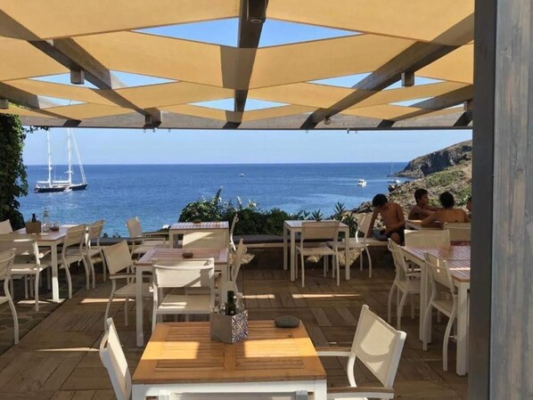 An outdoor seating area with tables and chairs, overlooking the sea with boats visible in the distance.