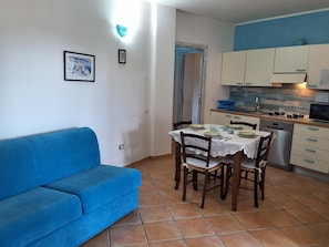 Een knus woongedeelte met een blauwe bank, een eettafel met stoelen, een kitchenette en een deur naar een andere kamer.