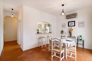 A dining area with a round table, white chairs, a hanging light fixture, and a wall-mounted air conditioner. - Apartment 'Medusa' close to the Sea with Balcony & Wi-Fi (OLHOS D´AGUA)