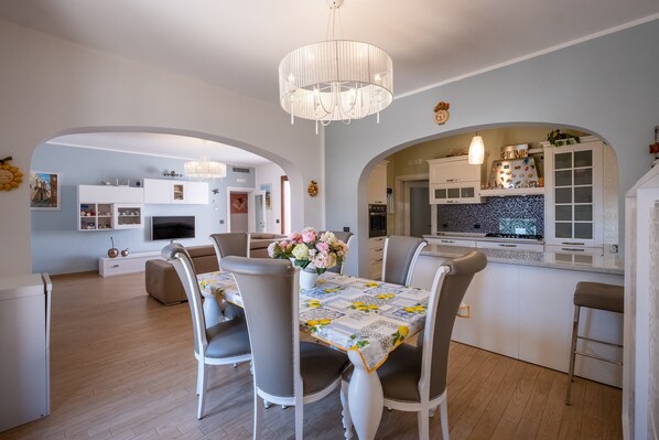 Une cuisine moderne avec des meubles blancs, un coin repas avec une table dressée pour quatre personnes, et un salon avec un canapé et un téléviseur.