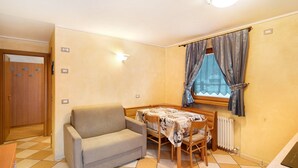 Ein kompakter Raum mit Schlafsofa, kleinem Tisch, Fenster mit Vorhängen, Heizkörper und Tür.
