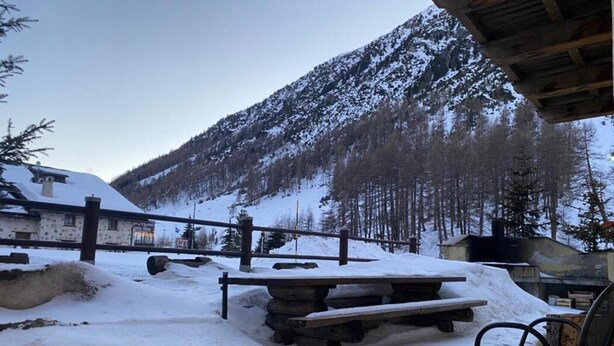 A snow-covered outdoor area with a wooden bench and a chair, a stone table, and a building with a snow-covered roof in the background. - Apartment "Piz Orsera" with Mountain View, Garden & Wi-Fi (Livigno)