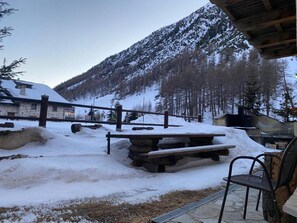 Un espace extérieur enneigé avec un banc en bois et une chaise, une table en pierre, et un bâtiment dont le toit est recouvert de neige, visible au fond.