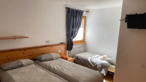 A room with two beds, a window with curtains, a wooden headboard, a radiator, and a wall-mounted TV. - Apartment "Piz Orsera" with Mountain View, Garden & Wi-Fi (Livigno)