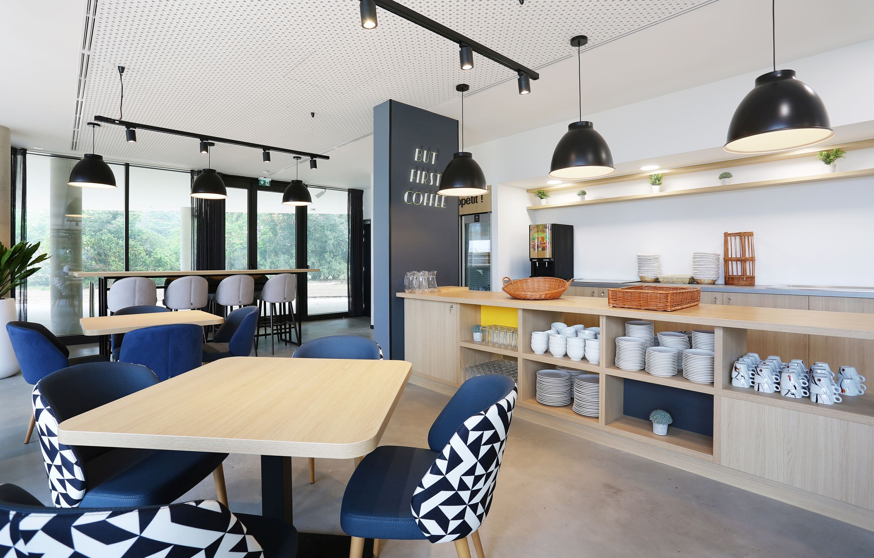 A modern cafe with a wooden table and blue chairs, a counter with various items, and a large window with a view of trees.