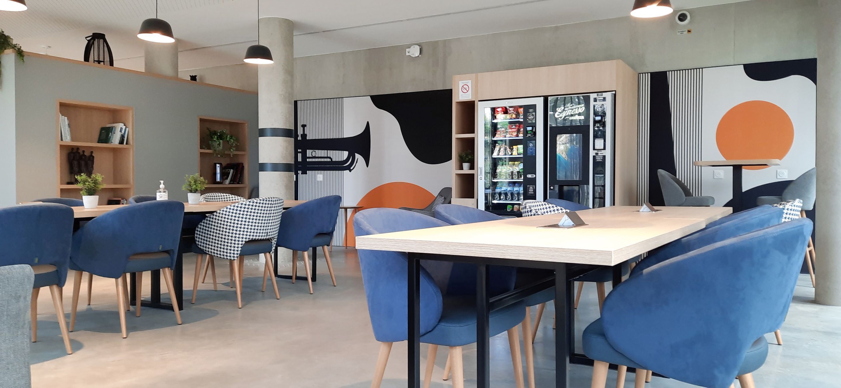 Un comedor moderno con sillas azules y una mesa de madera, una máquina expendedora y una pared con arte abstracto.