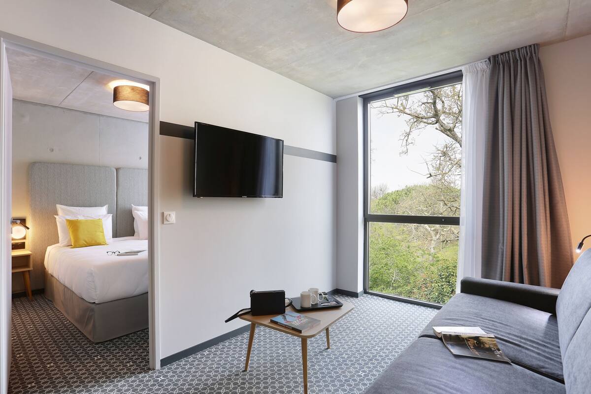 Ein Hotelzimmer mit einem Bett, einem an der Wand befestigten Fernseher, einem kleinen Tisch mit einem Buch und einer Tasse sowie einem großen Fenster mit Vorhängen.
