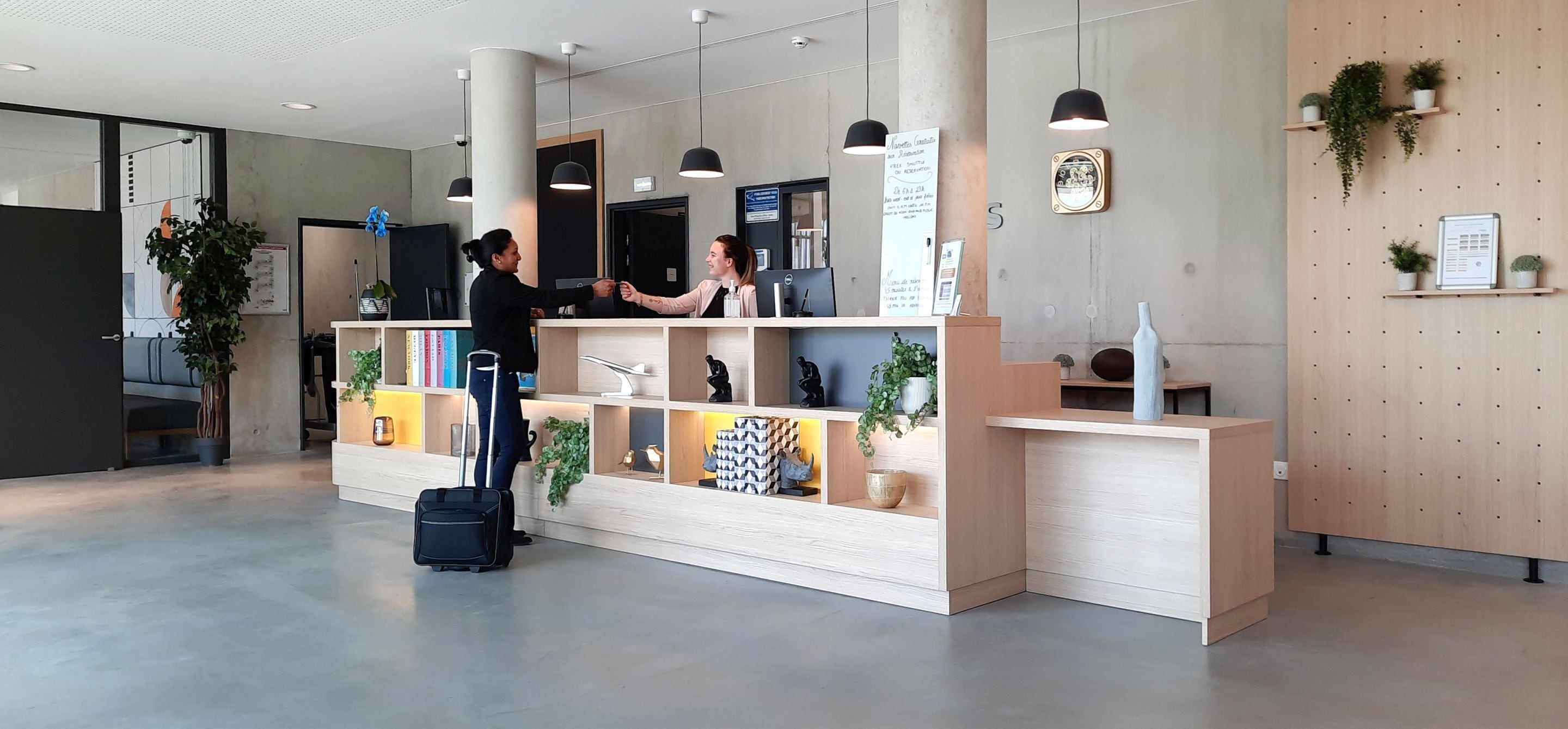 Un espace d’accueil avec un bureau en bois, des étagères ornées de plantes et une valise noire.