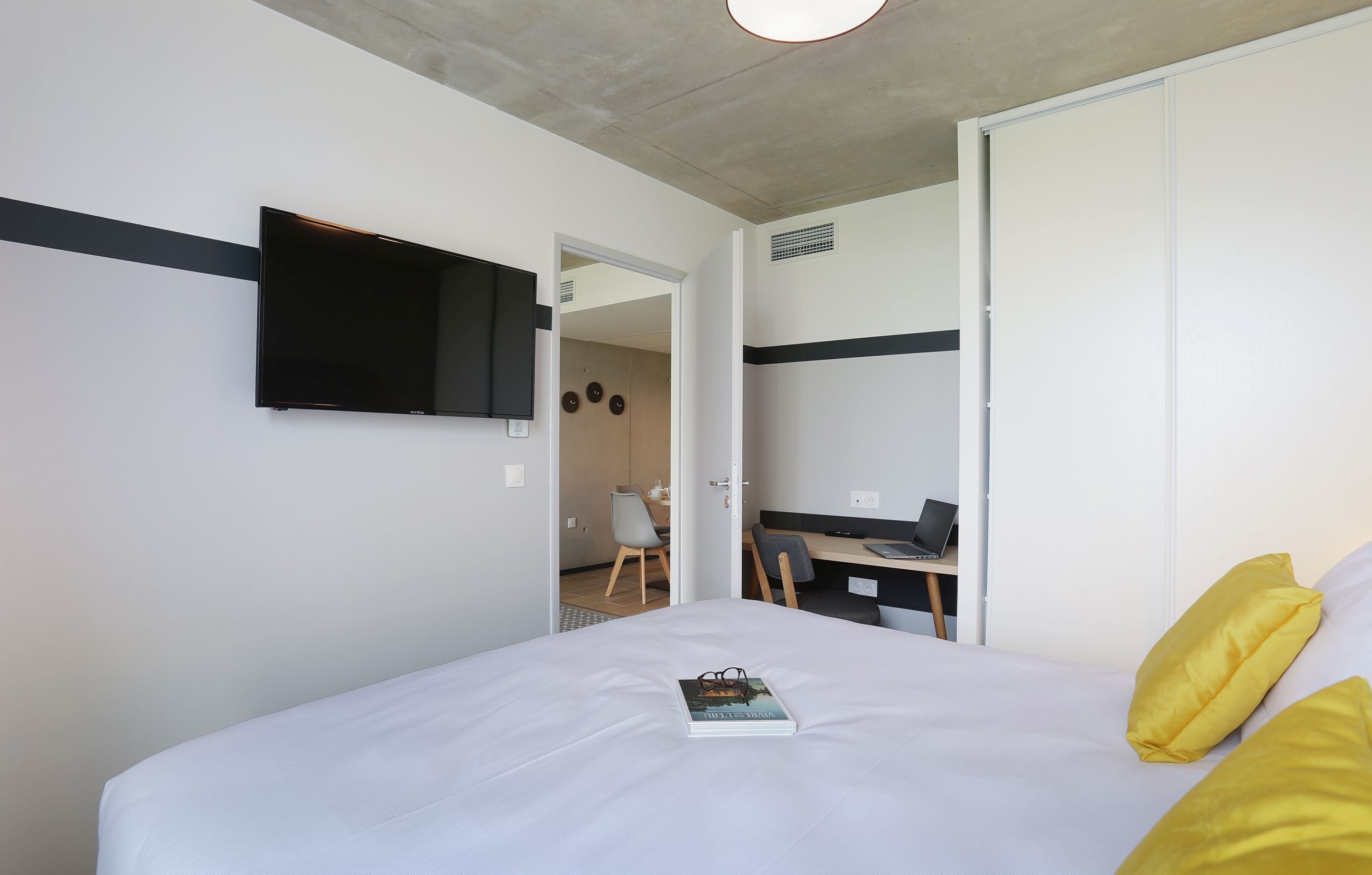 Una habitación de hotel moderna con una cama grande, un televisor de pantalla plana montado en la pared, un escritorio con una computadora portátil y una puerta que da a un baño.