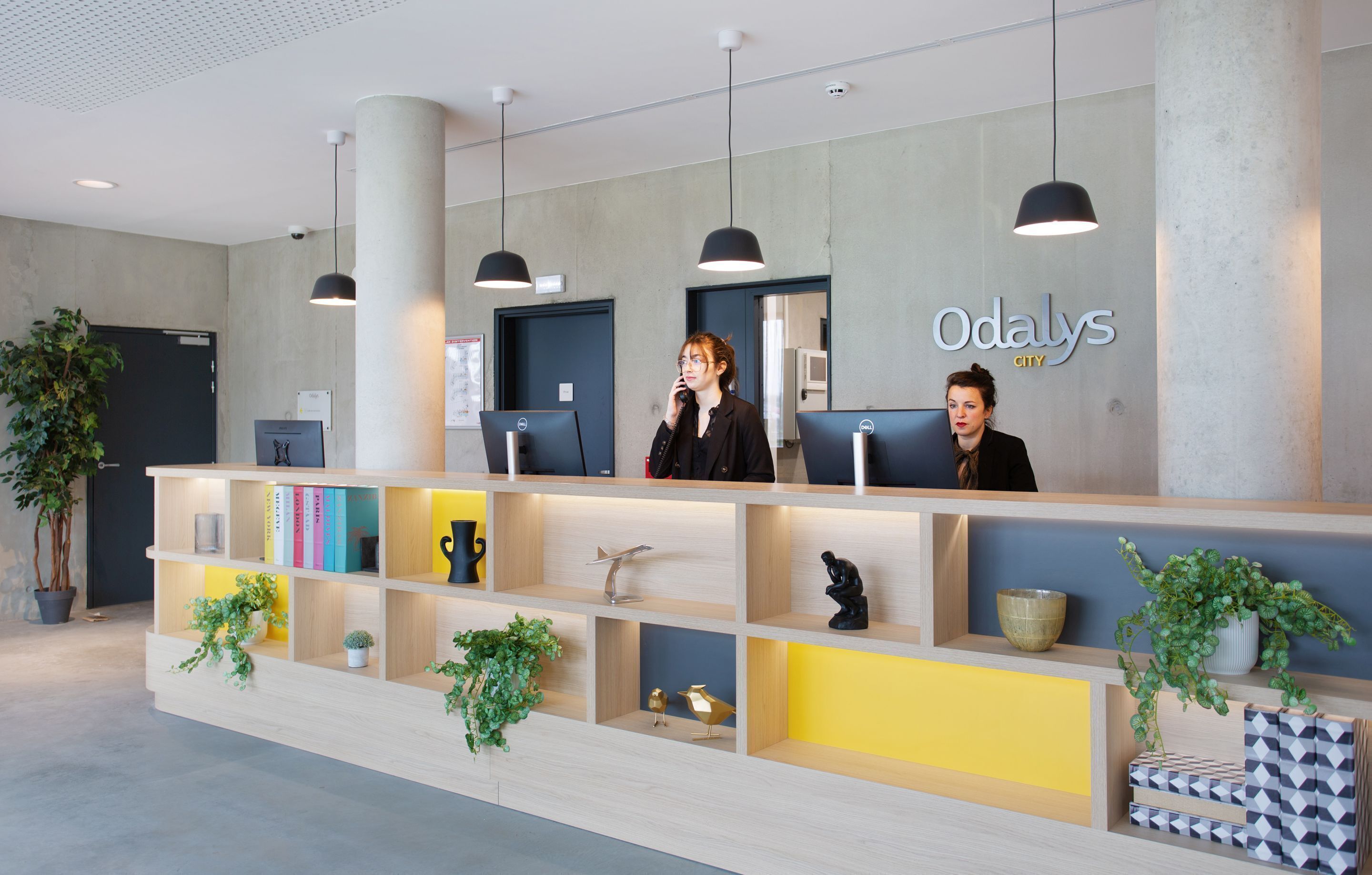 Un mostrador de recepción con tres monitores de computadora, un cartel que dice "Odalys City" y varios objetos decorativos, incluyendo plantas y figuritas.