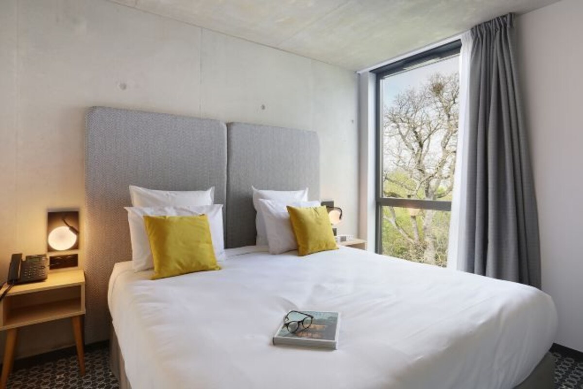 A hotel room with a large bed, two pillows, a bedside table with a lamp, and a window with curtains.