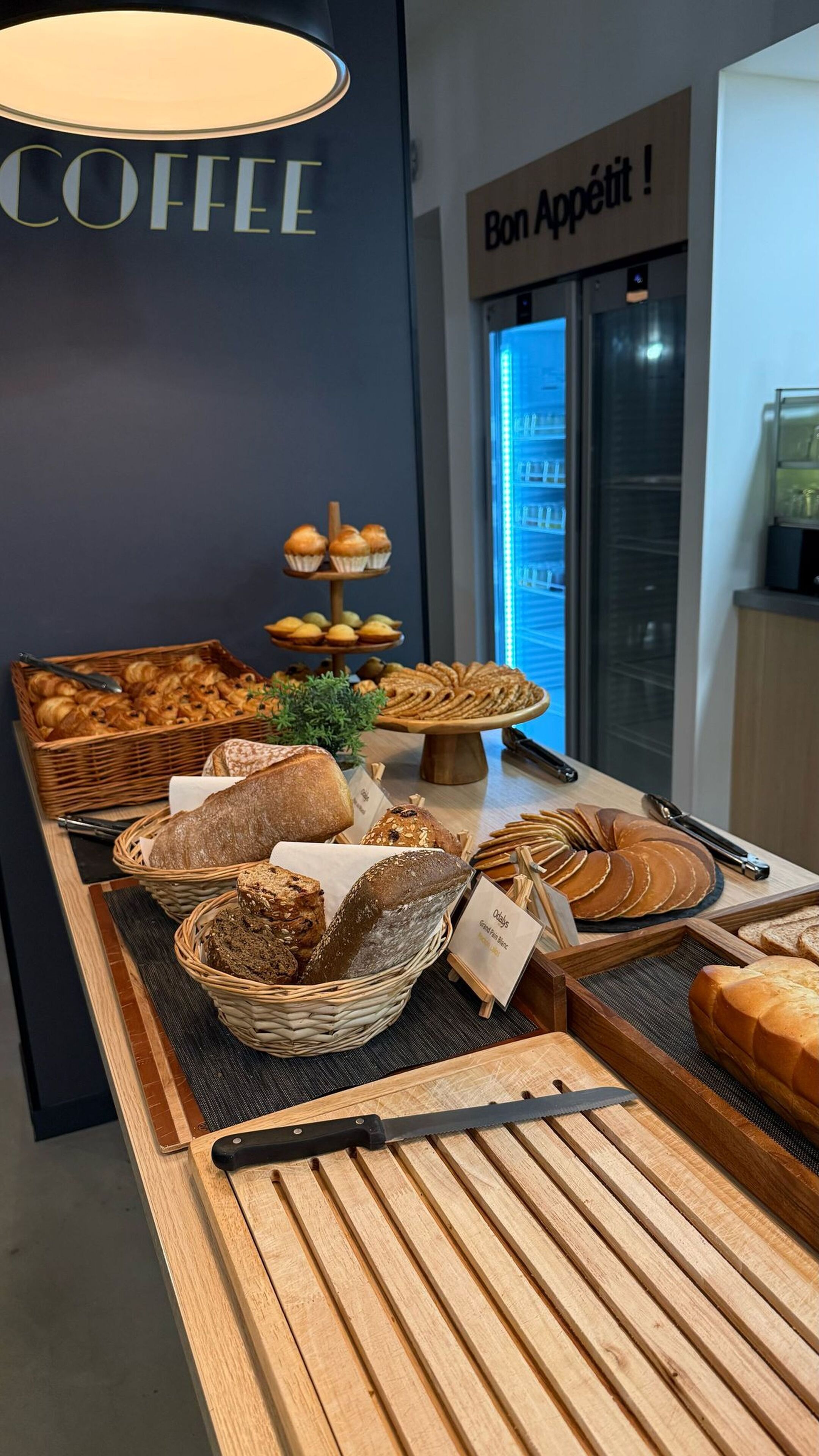 Mostrador de panadería con panes y pasteles variados, un cartel de café y un letrero que dice "Bon Appetit!".