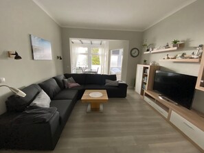 Un salon moderne avec un canapé d’angle noir, une table basse en bois, un téléviseur à écran plat posé sur un meuble en bois et une bibliothèque agrémentée d’objets décoratifs.