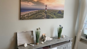 Eine Küstenlandschaftsmalerei mit Leuchtturm, Strand und Haus. Darunter befindet sich ein Regal mit Dekorationsobjekten, darunter eine Robbenfigur, eine Vase mit Gras und eine Muschel.