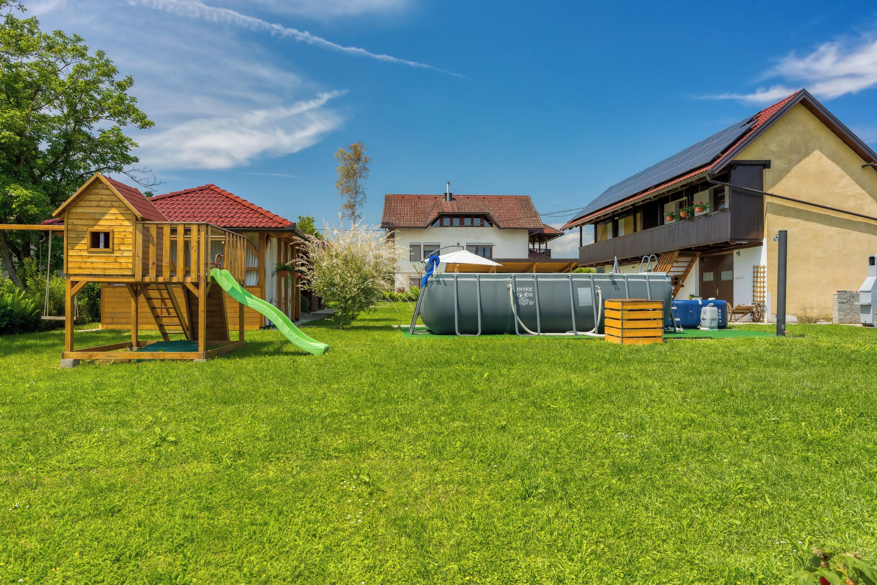 Una propiedad residencial con una casita de juegos de madera, un tobogán verde, un tanque de agua grande y una casa con techo rojo.