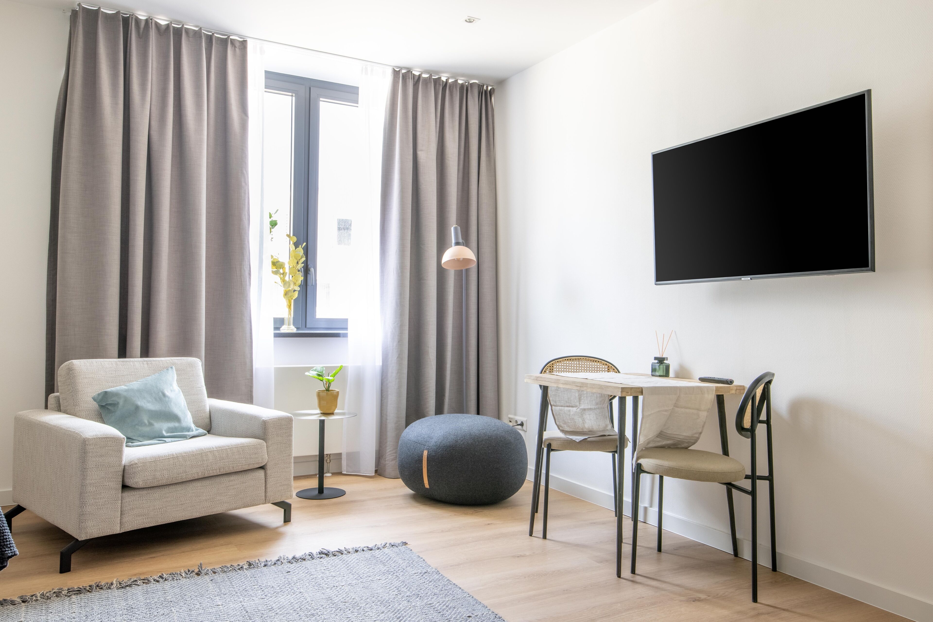 Una sala moderna con un sofá, un reposapiés redondo, una mesa de comedor con sillas, un televisor montado en la pared y una ventana con cortinas.
