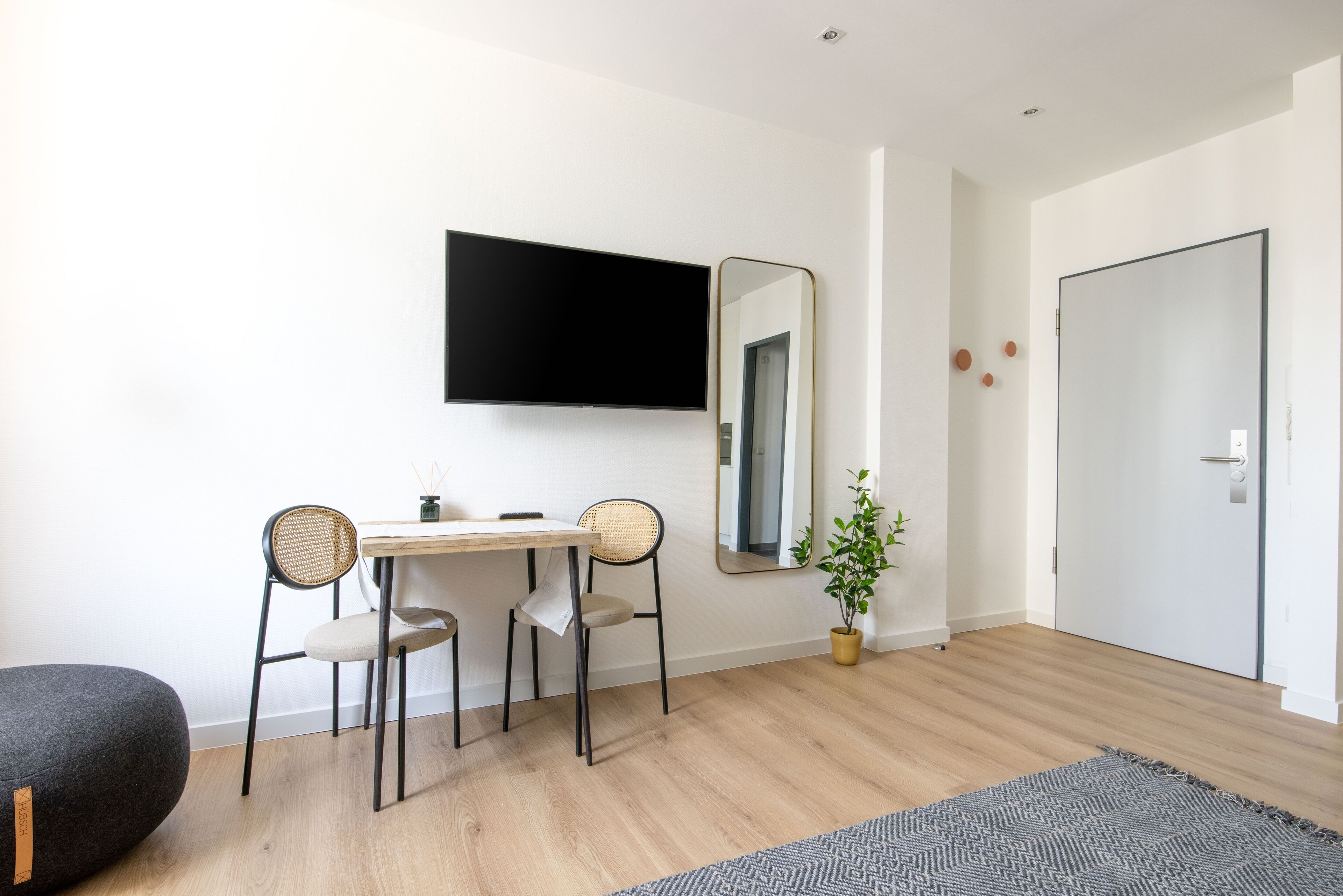 Una habitación moderna con piso de madera, un escritorio con dos sillas, un espejo grande, una planta en maceta y un otomano negro.