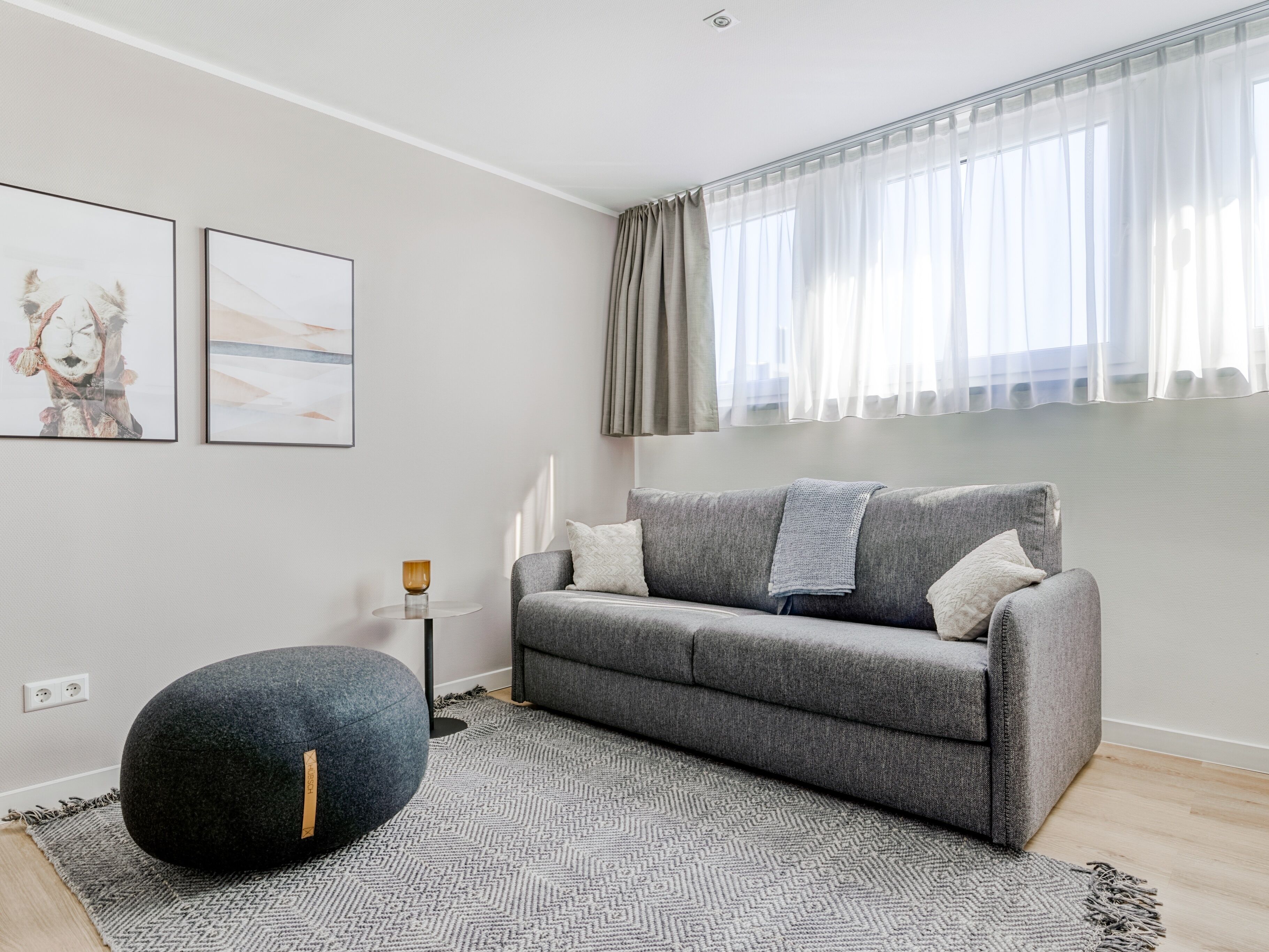 A modern living room with a grey sofa, a round black ottoman, and two framed artworks on the wall.