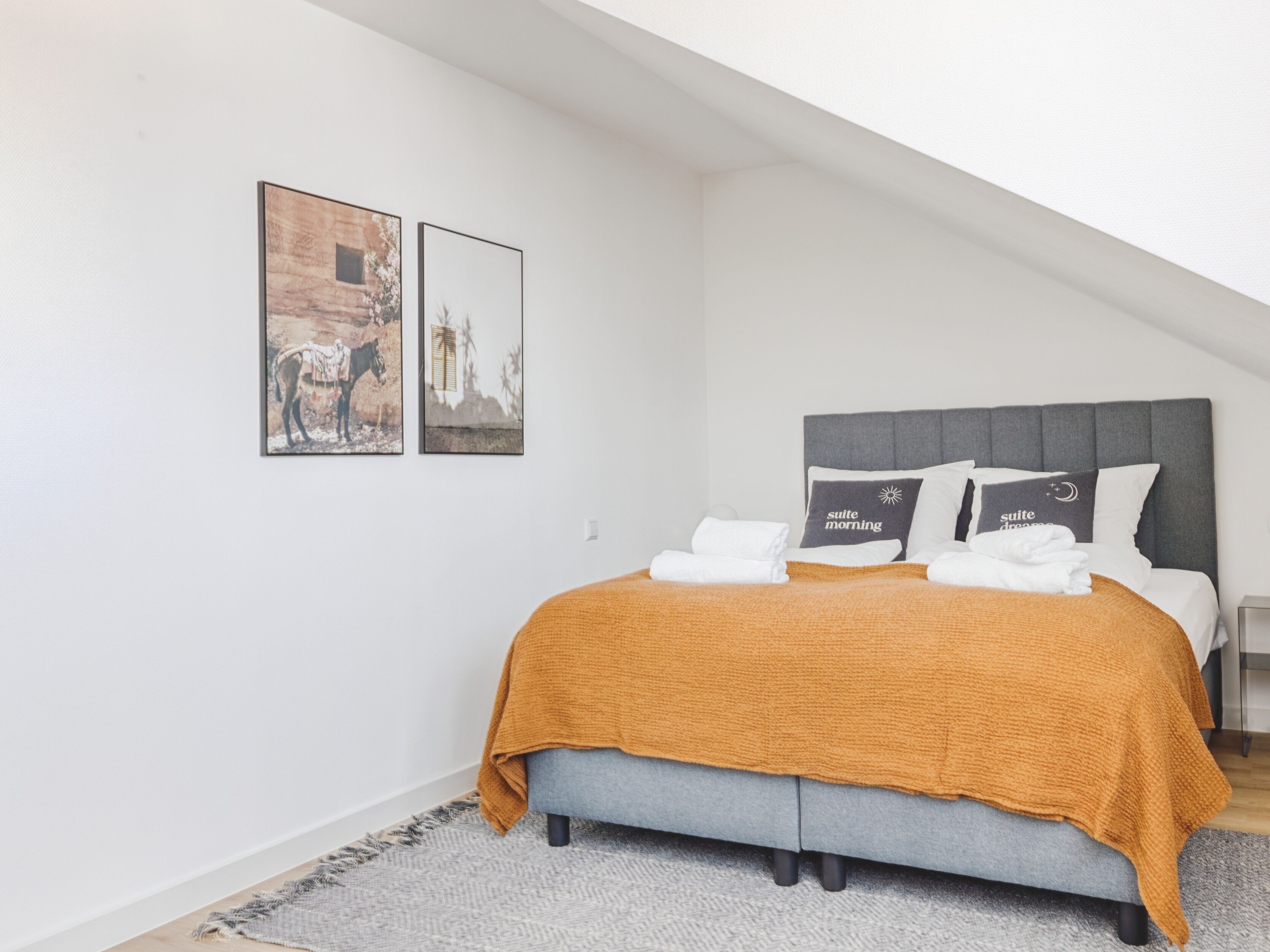A bedroom with a bed, pillows, and a blanket. There are two framed pictures on the wall.