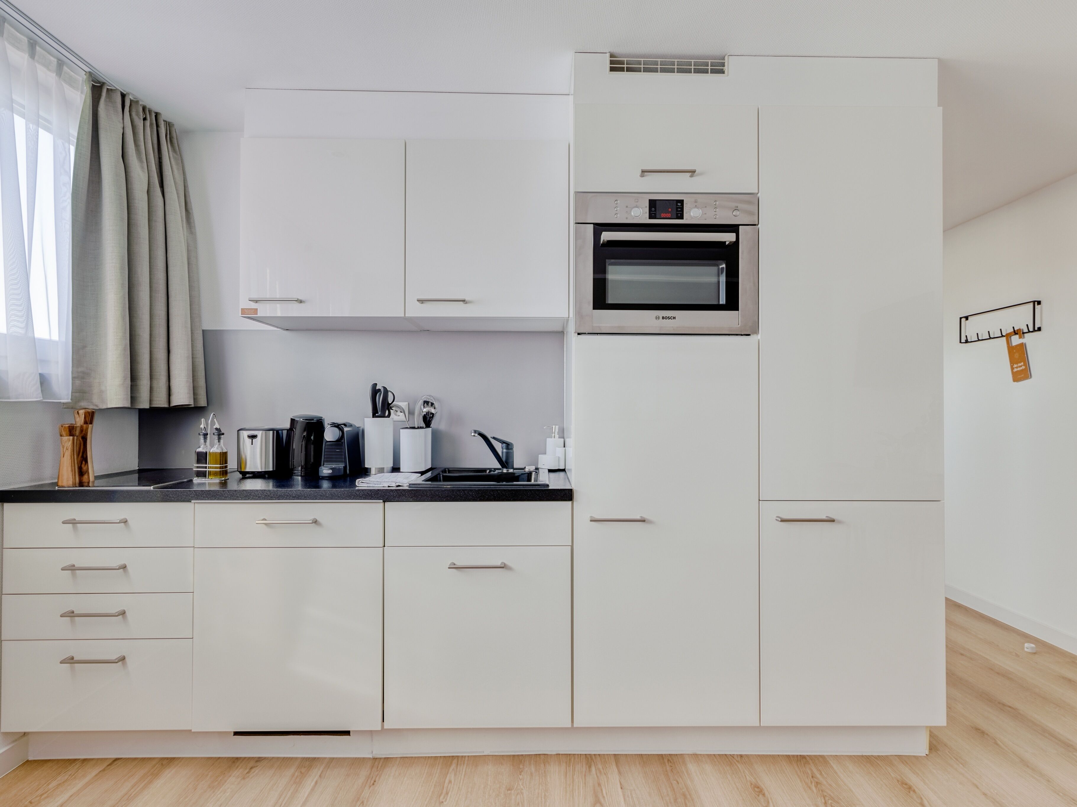 Una cucina moderna con mobili bianchi, un forno Bosch, un piano di lavoro nero e una finestra con tende leggere.