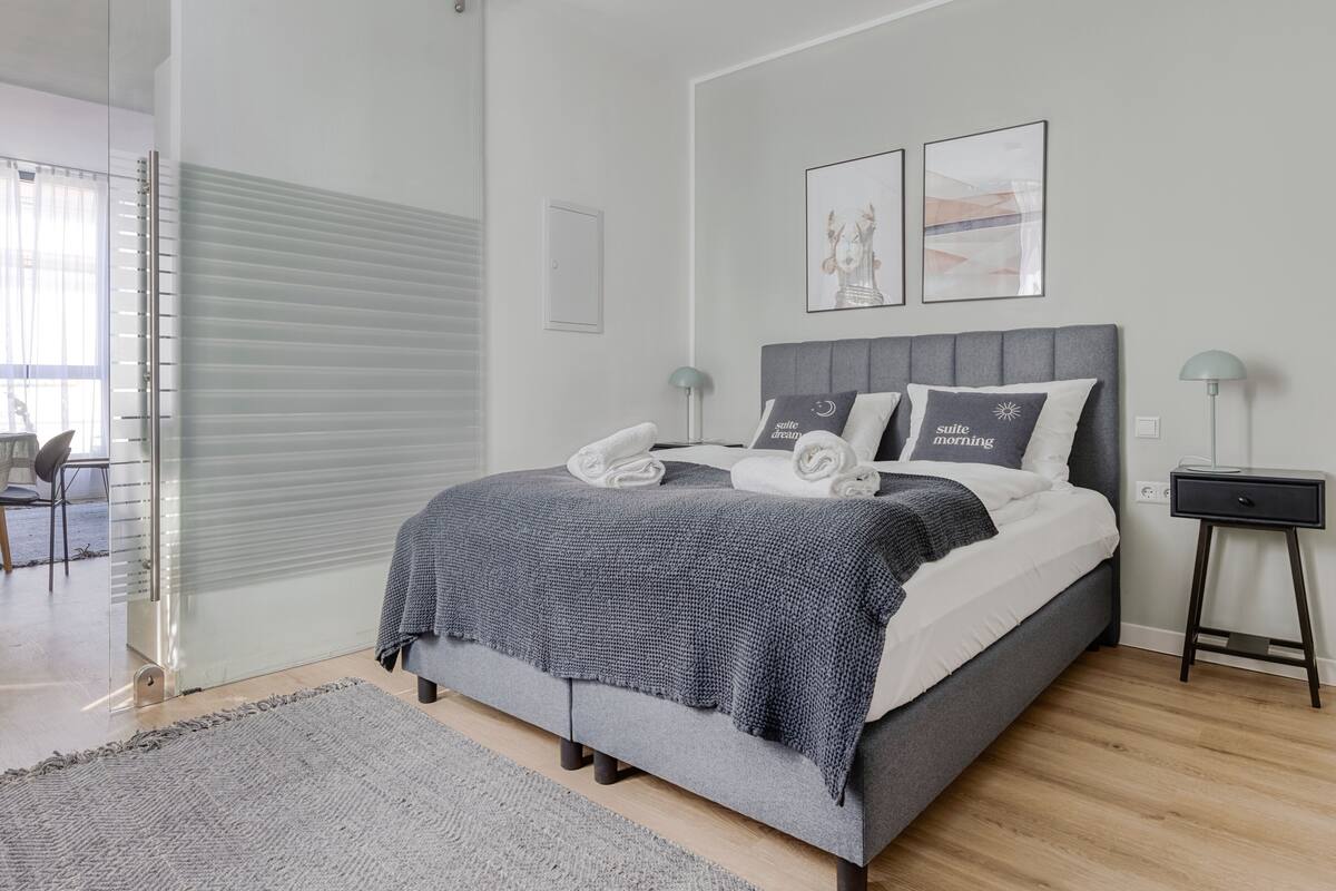 A modern bedroom with a grey bed, white pillows, and a grey blanket. There are two bedside tables with lamps, a grey rug, and a large window with blinds.