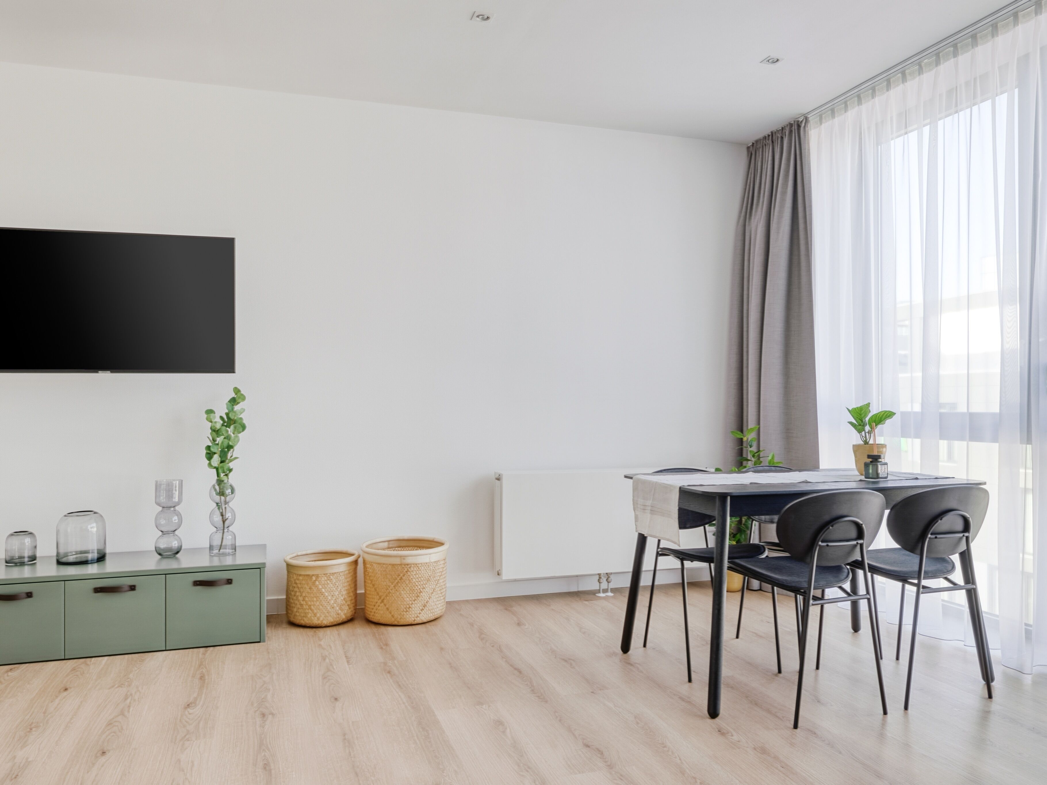 Una sala de estar moderna con un televisor de pantalla plana montado en la pared, una mesa de comedor con sillas, un radiador y dos cestos de mimbre en el suelo.