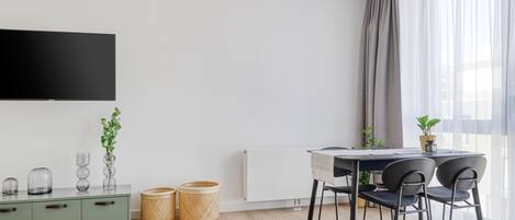 Una sala de estar moderna con un televisor de pantalla plana montado en la pared, una mesa de comedor con sillas, un radiador y dos cestos de mimbre en el suelo.