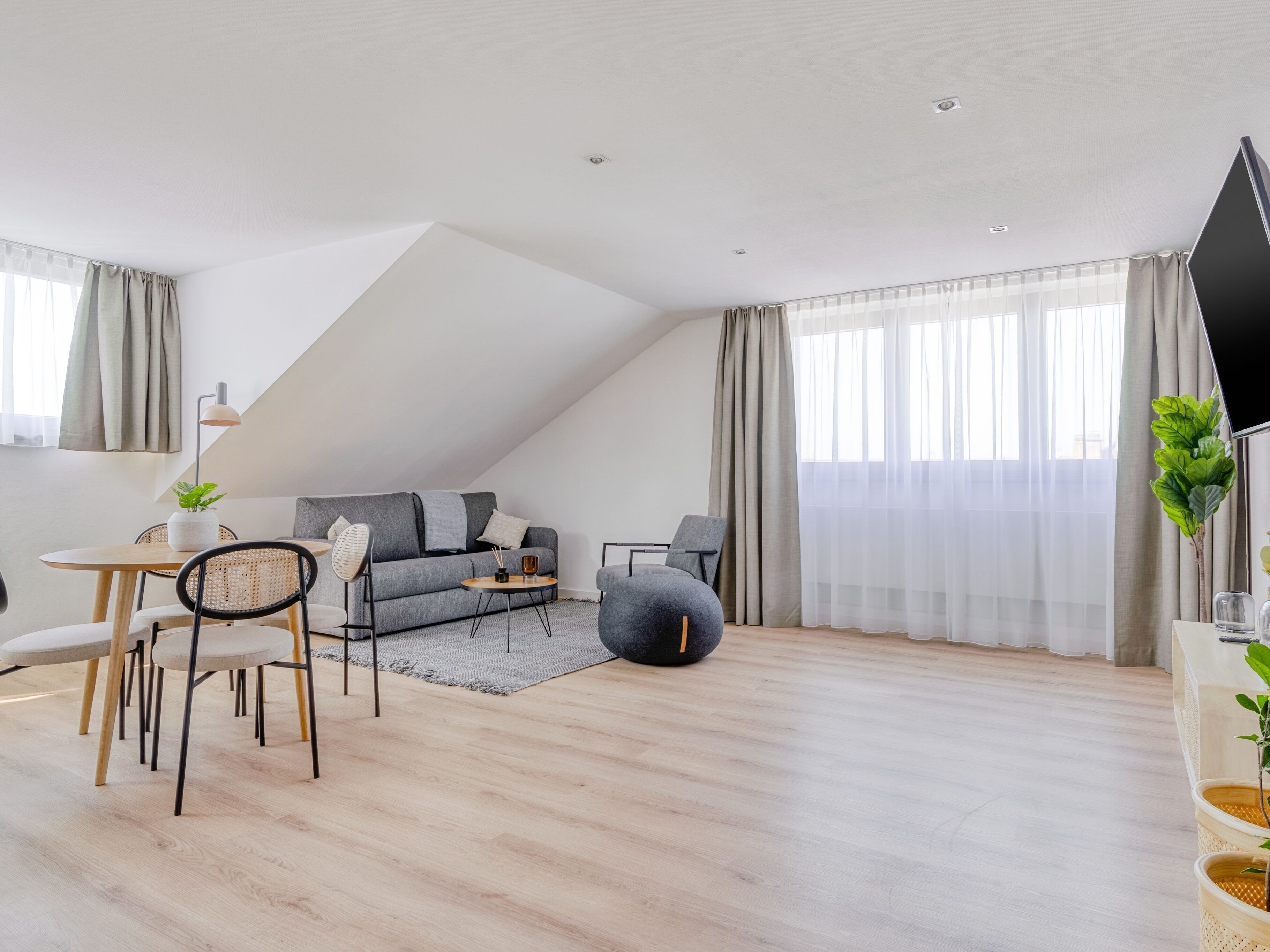 Un apartamento tipo loft moderno con zona de comedor, un sofá gris, un reposapiés negro y un gran ventanal con cortinas translúcidas.
