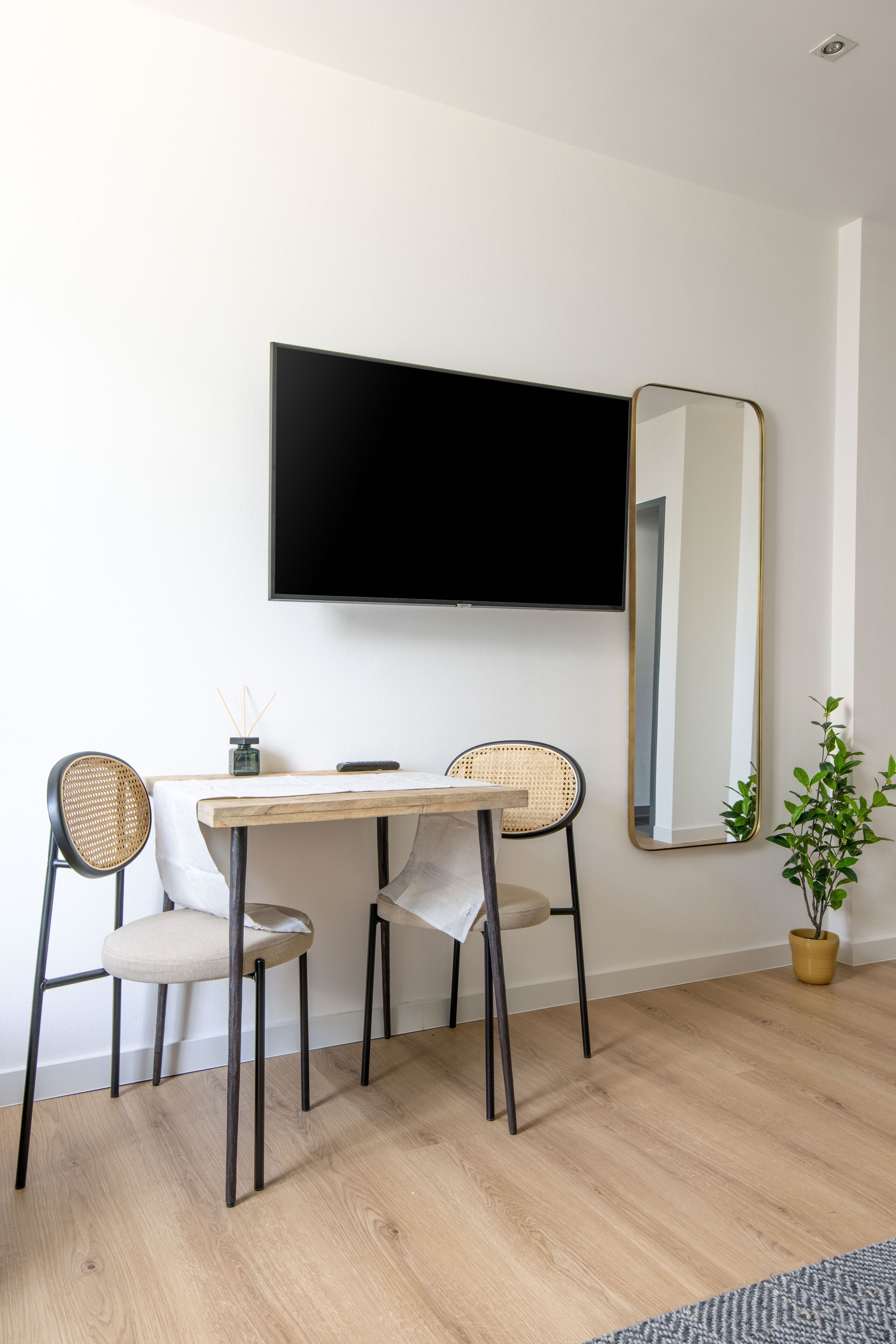 A modern room with a wooden dining table, two chairs, a flat-screen TV, a large mirror, and a potted plant.