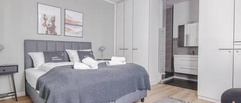 Un dormitorio moderno con una cama gris, un armario blanco y un baño visible a través de una puerta abierta.